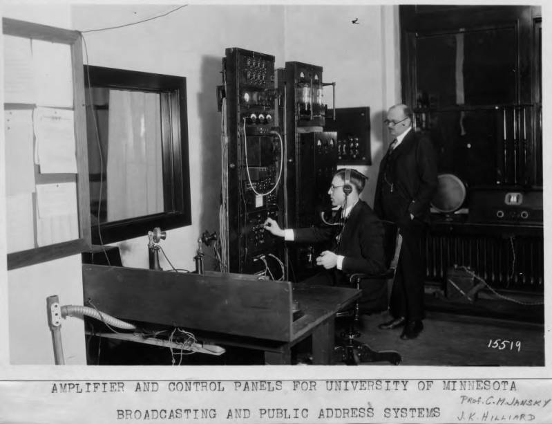 Black and white photo of two men wearing suits in a radio broadcast control room. One is seated and adjusting a dial. The other stands behind him with his hands in his pockets.