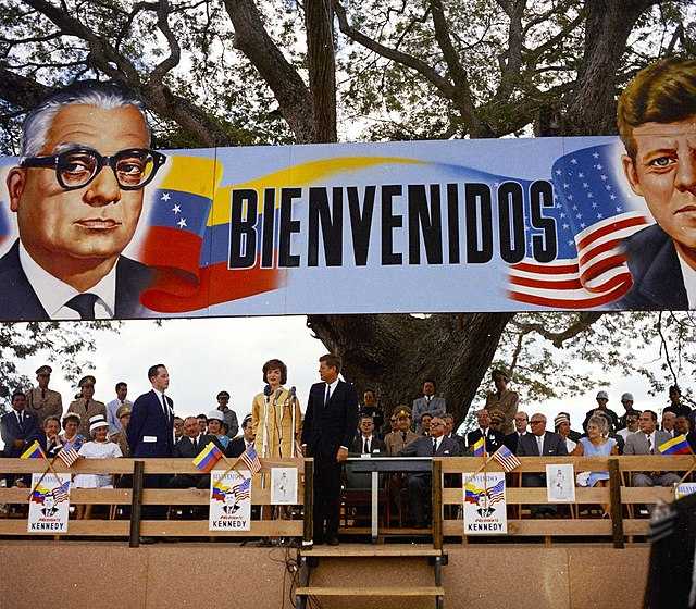 First Lady Jacqueline Kennedy delivering a speech in Spanish about the Agrarian Reform Program in La Morita, Venezuela, in 1961. Others pictured include President John F. Kennedy, President of Venezuela Rómulo Betancourt, and US Ambassador to Venezuela Teodoro Moscoso. Public domain photograph taken by Cecil Stoughton, retrieved via [Wikimedia](https://commons.wikimedia.org/wiki/File:Alliance_for_Progress_in_Venezuela_1961.jpg).